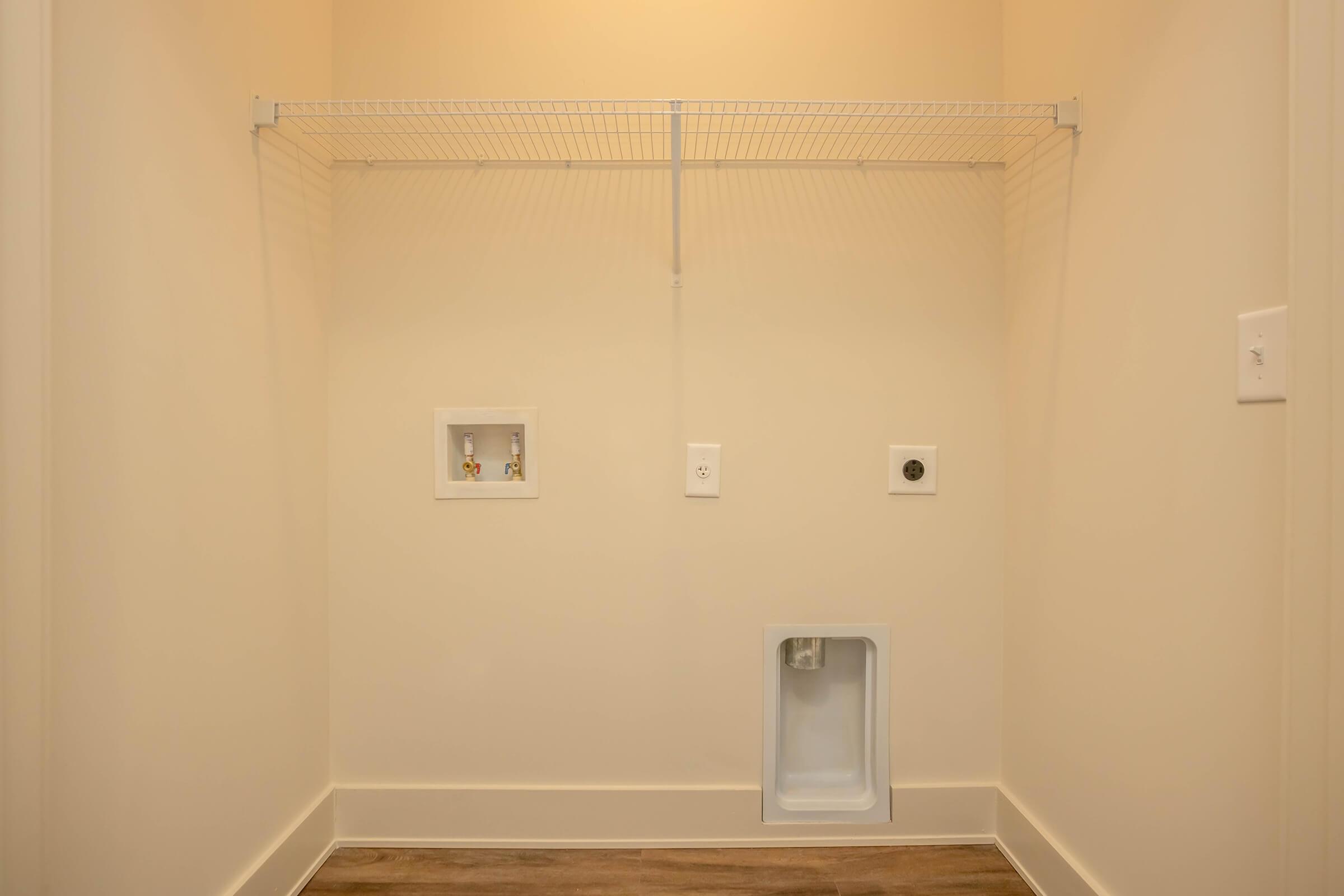 A spacious laundry room with light-colored walls and a wood-style floor. It features a wire shelf for storage, two electrical outlets, and a water connection, with a small opening at the bottom for pet access or venting. The overall space is clean and empty, ready for appliances.