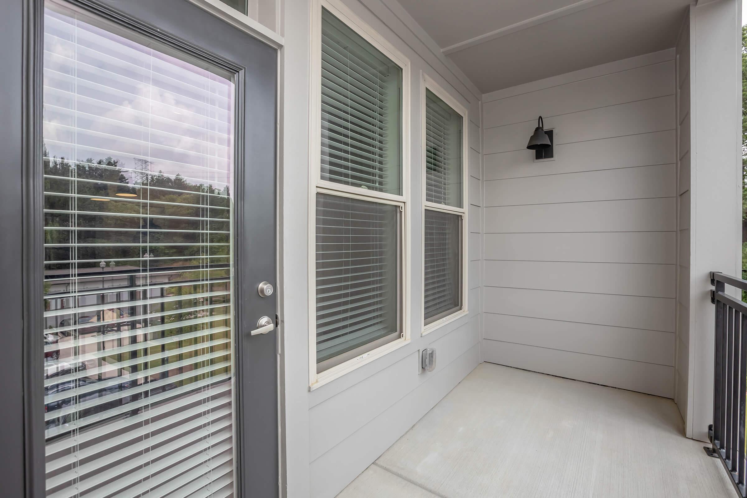 A small balcony or porch area with light gray walls, featuring a door with glass panels and blinds. There is a light fixture mounted on the wall, and the floor is a light, smooth surface. The view shows a hint of greenery through the windows.