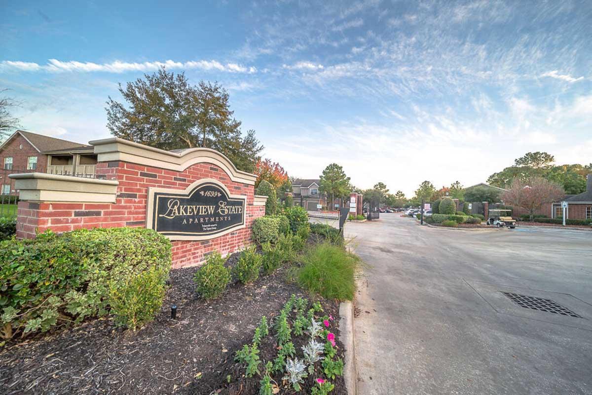 A view of the entrance to Arkview State Apartments, featuring a brick sign with the name prominently displayed. Surrounding the sign are landscaped areas with flowers and greenery. In the background, there are residential buildings and a clear sky.