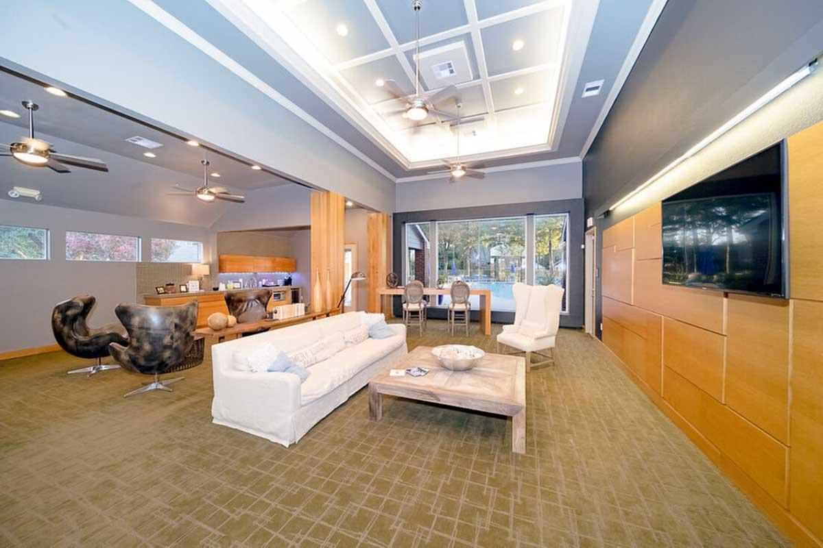 A modern living room featuring a light-colored sofa, a wooden coffee table, and stylish armchairs. Large windows provide natural light and a view of the outdoors. The room has a contemporary design with ceiling fans, a coffered ceiling, and a wall-mounted TV. Elegant decor creates a welcoming atmosphere.