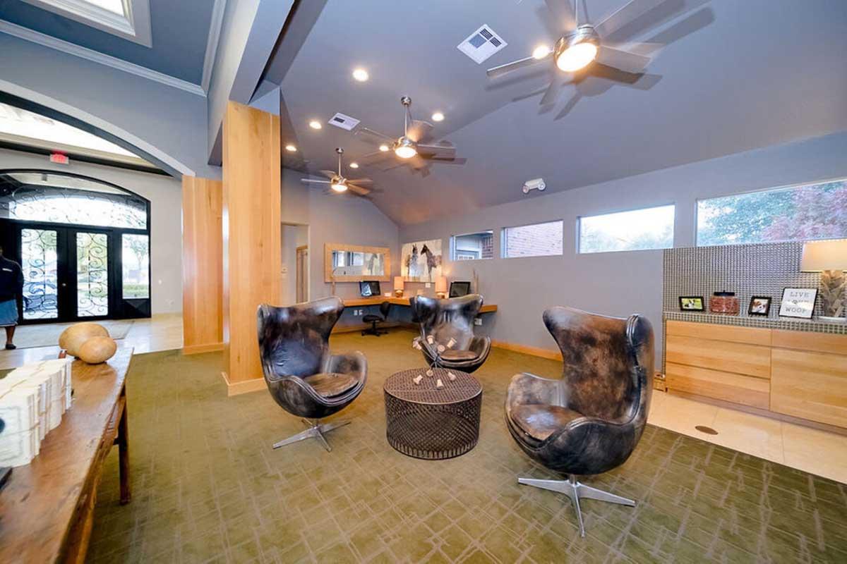 Modern interior of a lounge area featuring two stylish black chairs and a round table. The space has a high ceiling with ceiling fans, warm wood accents, and a green carpet. Light streams in through large windows, creating an inviting and contemporary atmosphere.