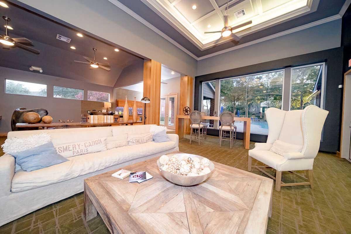 A modern living space featuring a light-colored sofa with decorative pillows, a wooden coffee table, and a cozy wingback chair. Large windows provide natural light and a view of the outdoors, while stylish ceiling fans and contemporary decor enhance the inviting atmosphere.