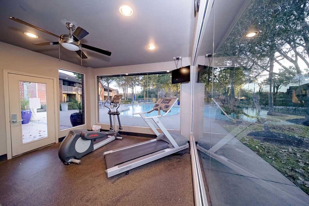 A bright fitness room featuring a treadmill and an exercise bike, with large windows overlooking a swimming pool and outdoor area. The space is well-lit with ceiling lights and has a modern aesthetic, complete with a ceiling fan for airflow.