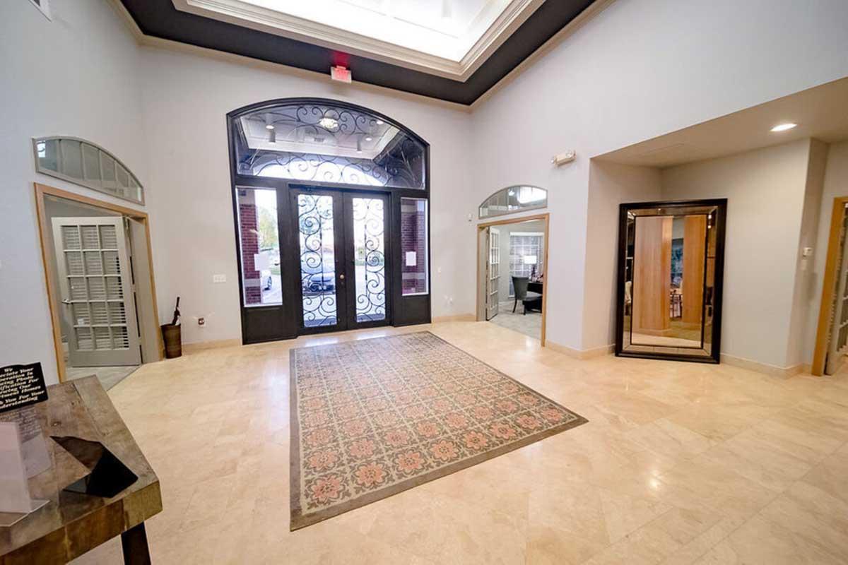 Spacious entryway featuring a decorative tile floor, large double doors with ornate ironwork, and high ceilings. The walls are painted in a neutral color, and the area is well-lit by natural light. To the right, there's a glass door leading to another room, adding to the open feel of the space.