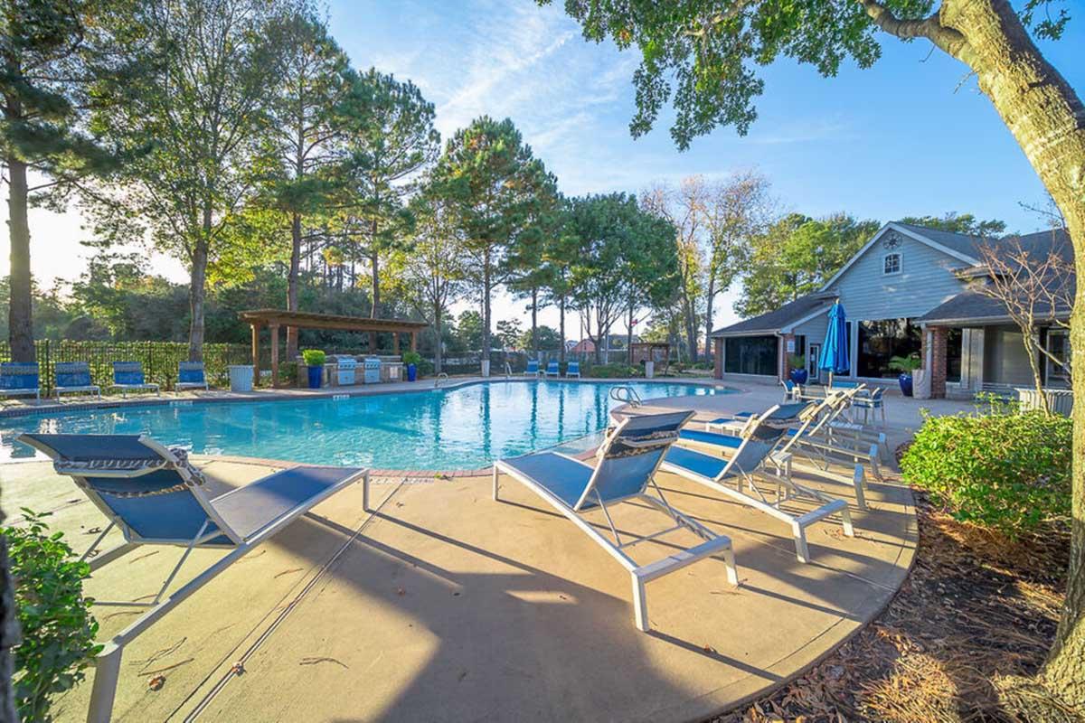 A serene outdoor swimming pool surrounded by lounge chairs, trees, and a small building. The pool area is well-maintained, with a sunny sky and green scenery, creating a relaxing atmosphere for leisure and gatherings.