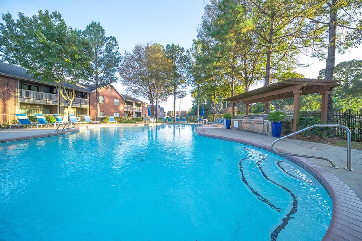 A clear swimming pool surrounded by green trees and lounge chairs. In the background, residential buildings can be seen with balconies overlooking the pool area. A shaded pergola with plants is located near the pool, creating a relaxing outdoor atmosphere.