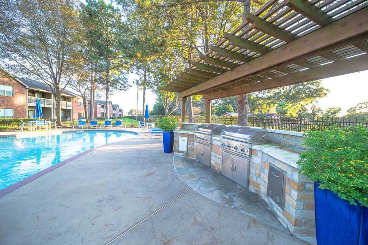 Outdoor swimming pool area with lounge chairs, surrounded by trees. To the left, there are several stainless steel grills under a wooden pergola, with potted plants nearby. The scene is bright, suggesting a pleasant day.