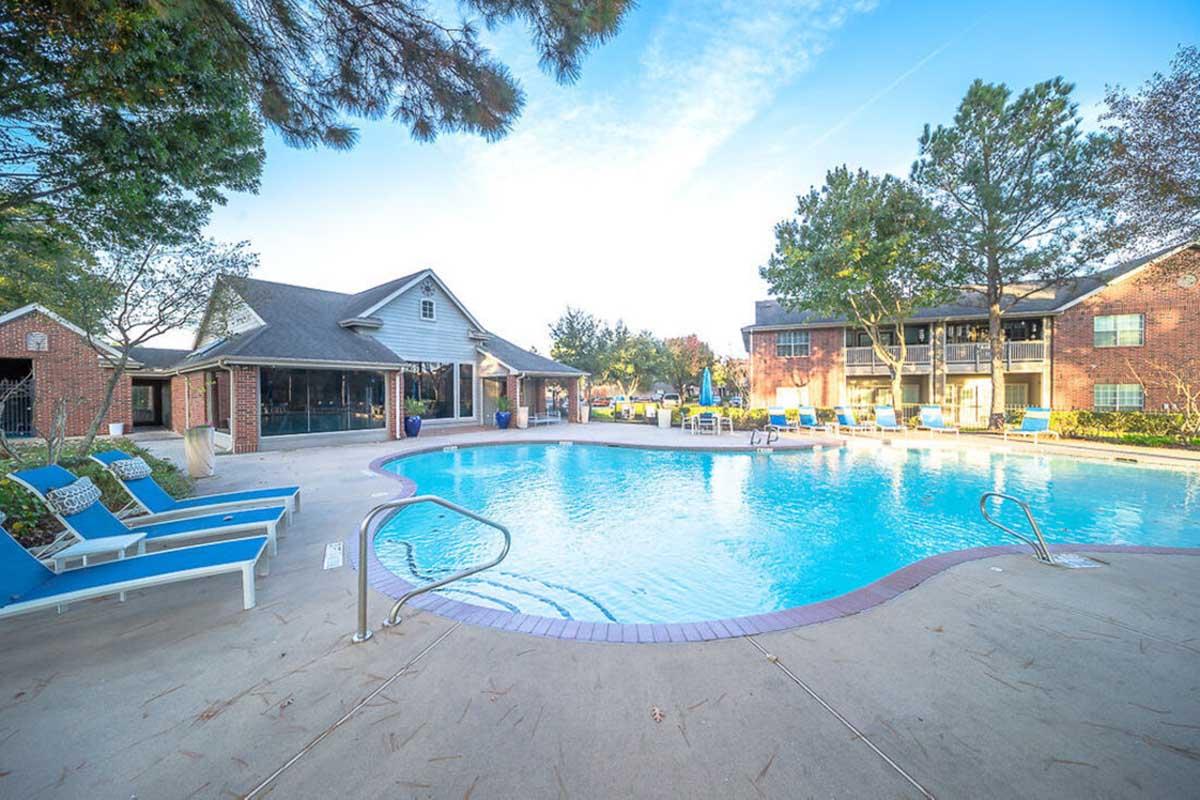 A clear blue swimming pool surrounded by lounge chairs and a few trees. In the background, there is a building with large windows, likely an apartment complex or clubhouse. The scene is bright and inviting, suggesting a relaxing atmosphere perfect for leisure activities.