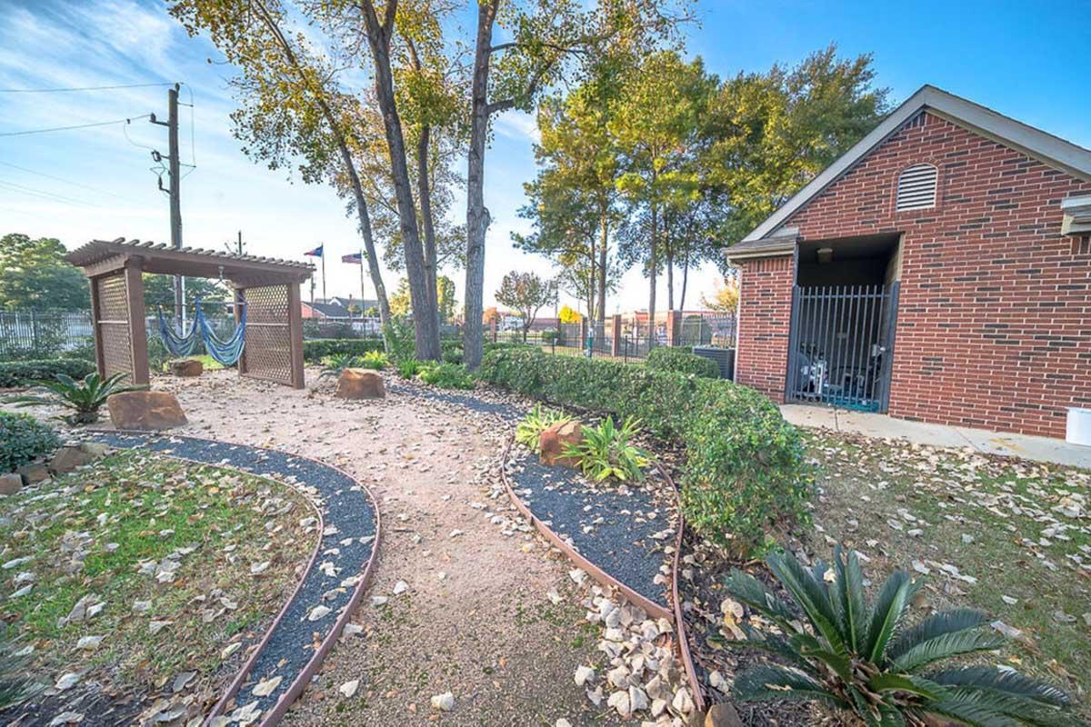 A pathway lined with stones winds through a landscaped area with shrubs and small plants. To the left, a wooden pergola supports a hammock, while to the right, a brick building features a gated entrance. The scene is set under a clear blue sky with autumn leaves scattered on the ground.