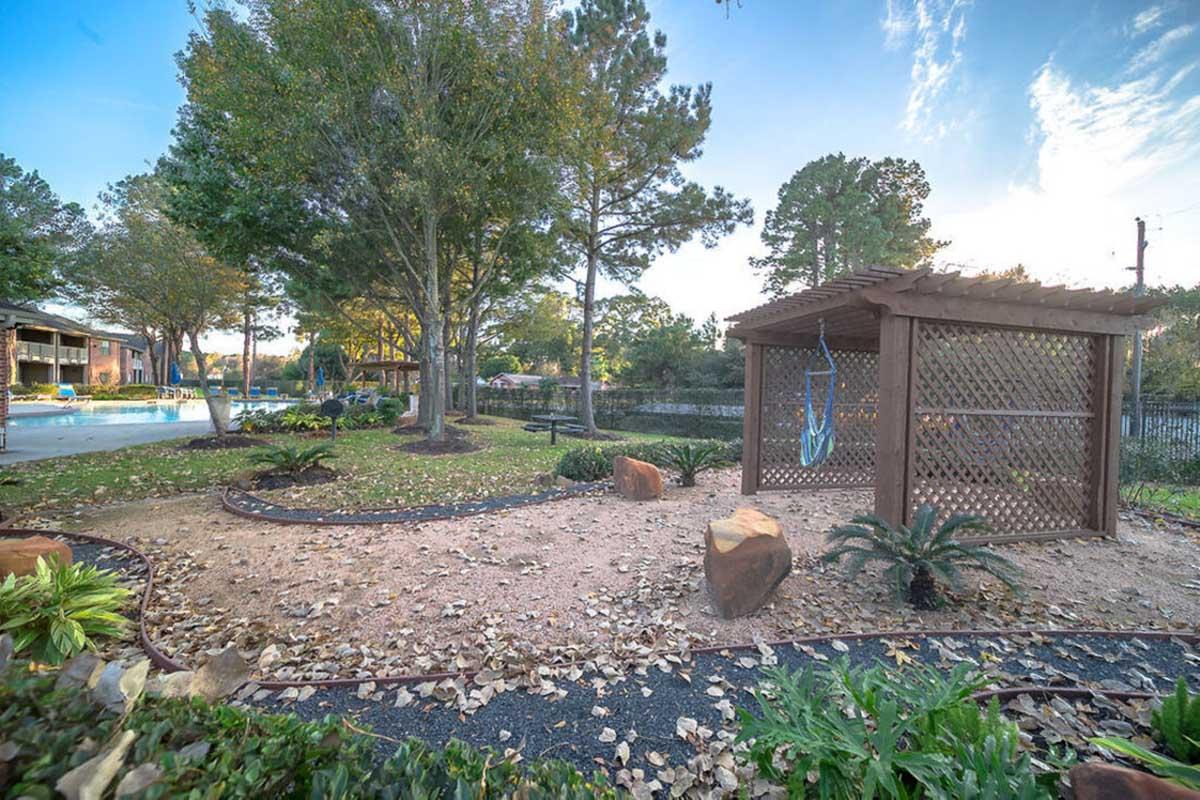 A serene garden scene featuring a shaded area with a wooden pergola, surrounded by lush greenery and decorative rocks. In the background, there is a pool area and a peaceful landscape with trees under a clear blue sky. Dry leaves are scattered on the ground, adding to the tranquil atmosphere.
