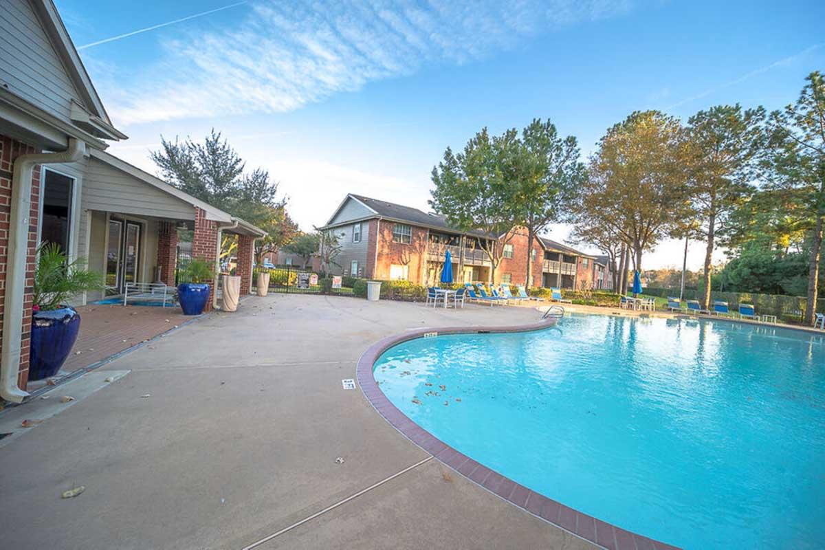 A serene swimming pool at an apartment complex, surrounded by residential buildings and lush trees. The pool area features a curved deck with lounge chairs and vibrant blue planters. The sky is clear with wispy clouds, creating a tranquil atmosphere for relaxation.