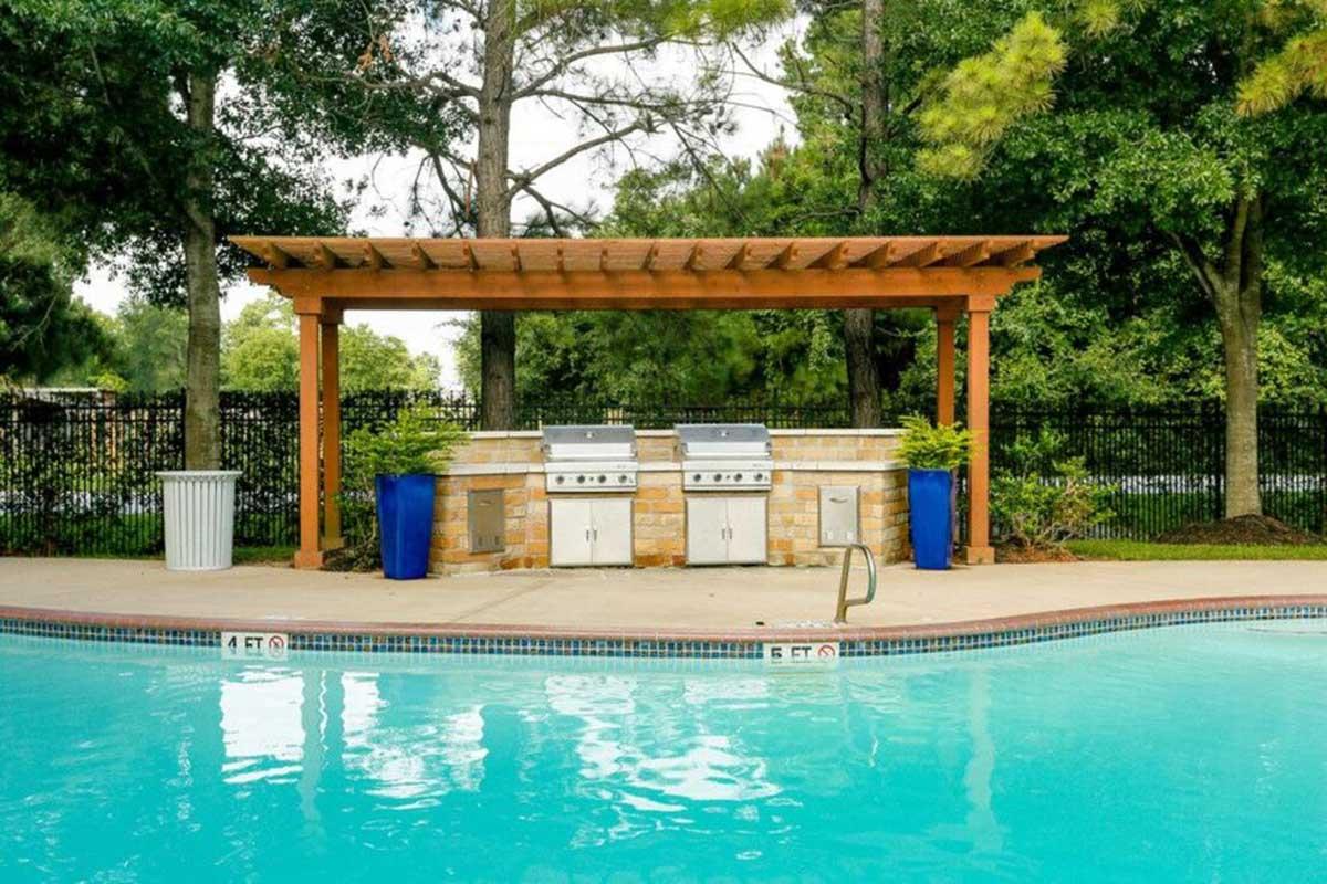 A tranquil swimming pool area featuring a wooden pergola with two grills underneath, surrounded by lush greenery. There are blue planters and a waste bin nearby, creating a relaxing outdoor space for gathering and cooking by the pool.