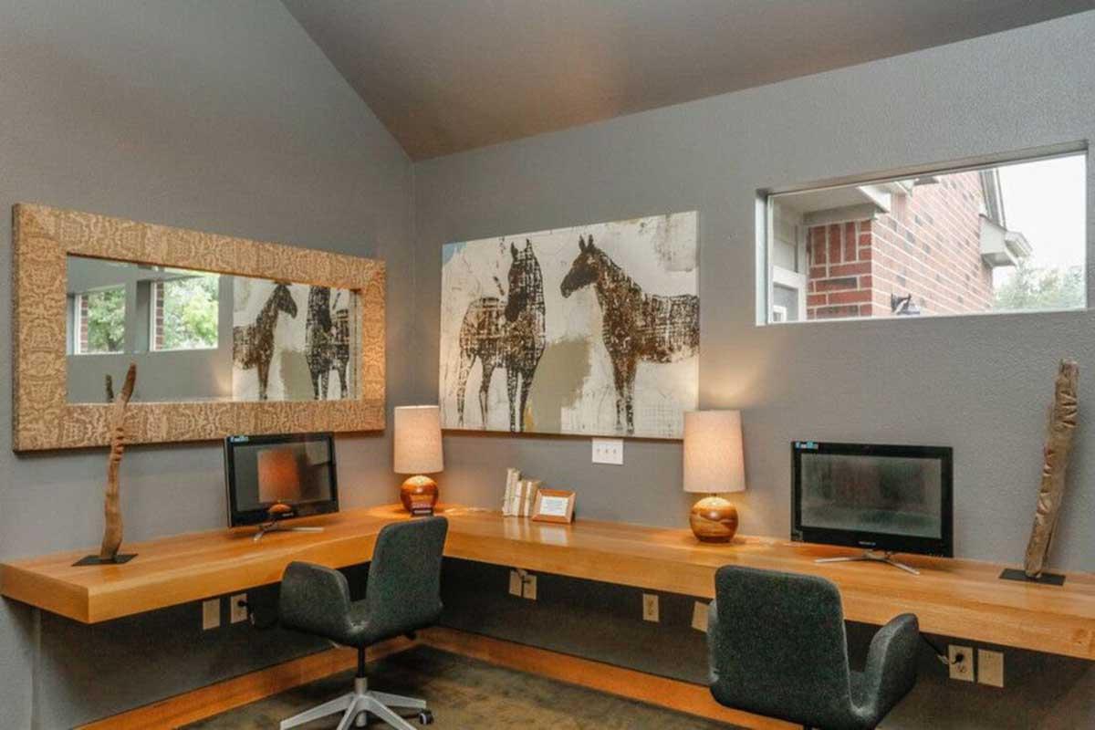 A modern workspace featuring two wooden desks with black chairs, two computers, and decorative items. The walls are painted gray and adorned with a large painting of horses and a decorative mirror. Natural light filters in through a window, creating a bright and inviting atmosphere.