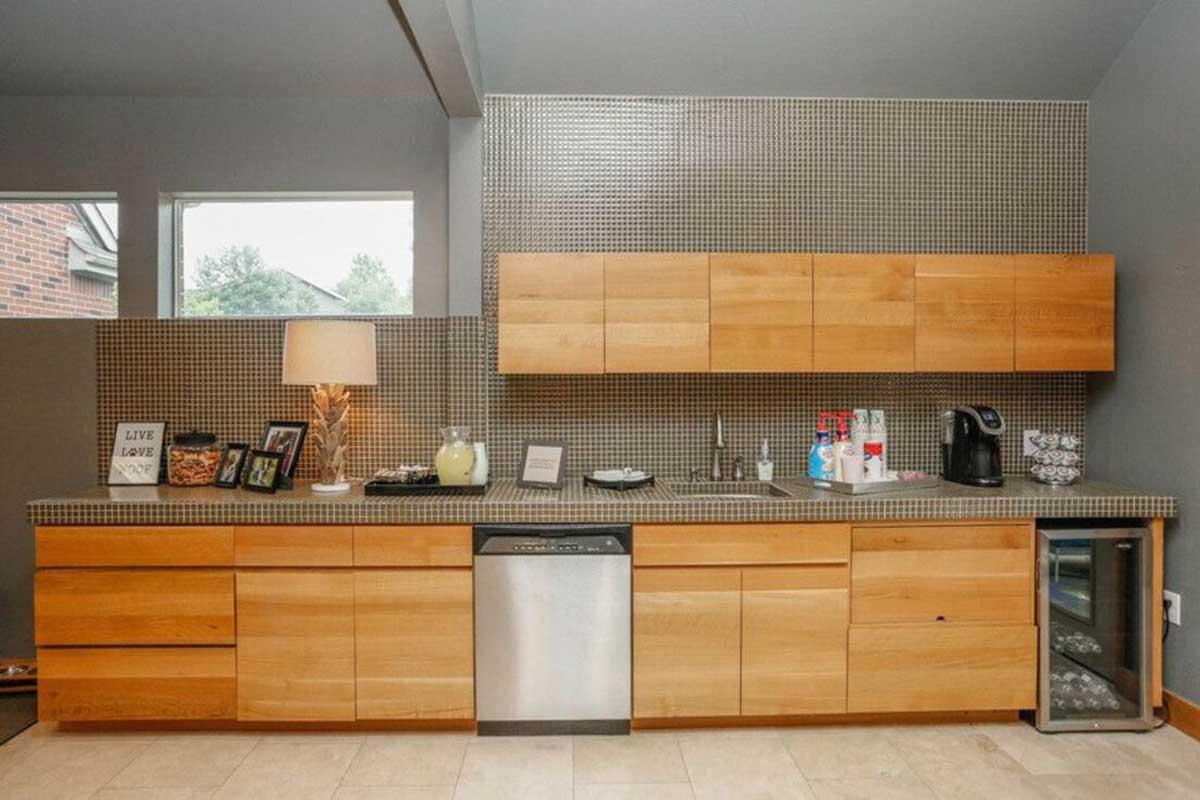 Modern kitchen featuring wooden cabinets with a sleek design, a stainless steel dishwasher, and a countertop with various appliances including a coffee maker and a beverage fridge. The space is well-lit with large windows, and a lamp sits on the counter beside framed photos.
