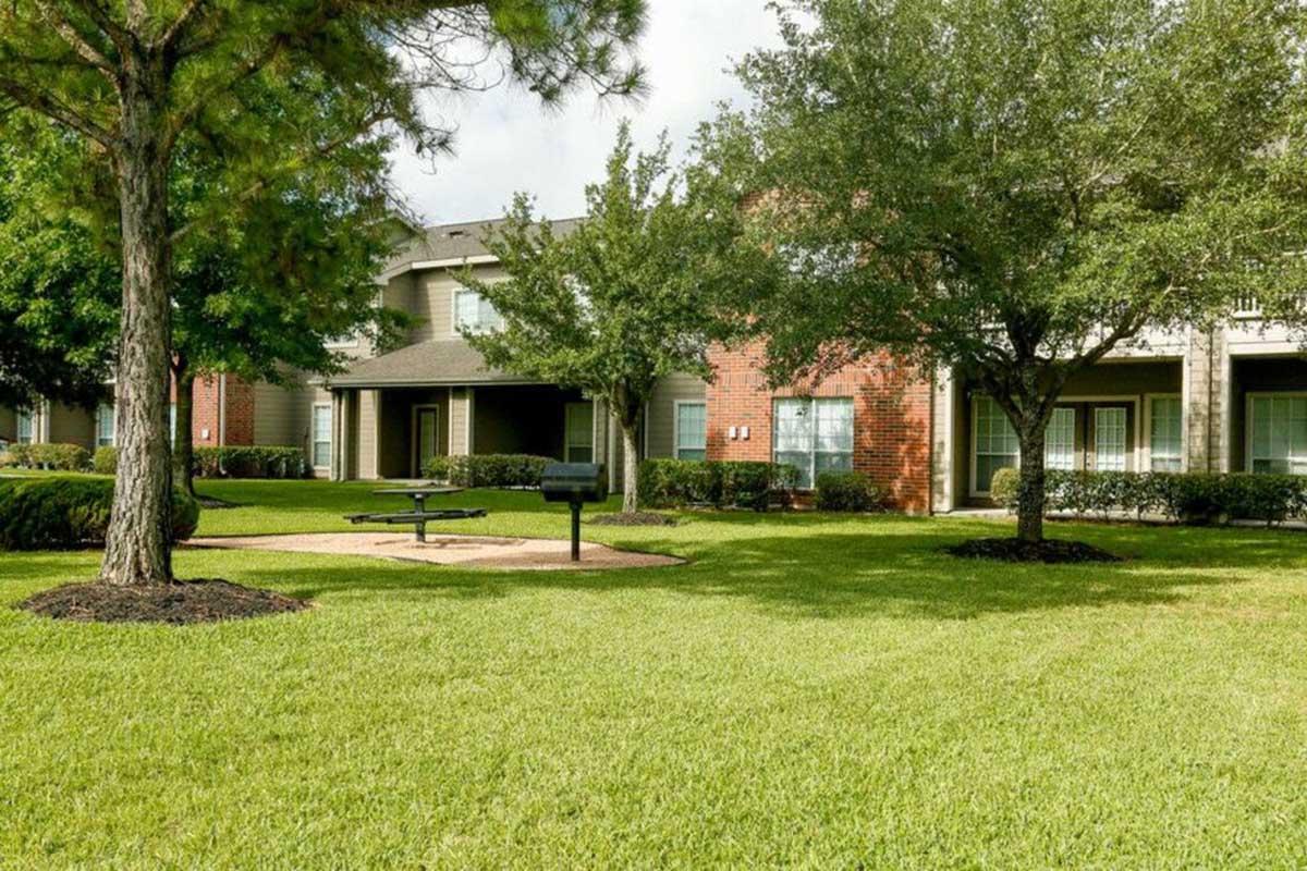 A well-maintained apartment complex surrounded by lush green grass and trees. Features include a picnic area with a bench and grill, and buildings with red brick and light siding in the background. The scene is bright and open, showcasing a peaceful outdoor space for residents.