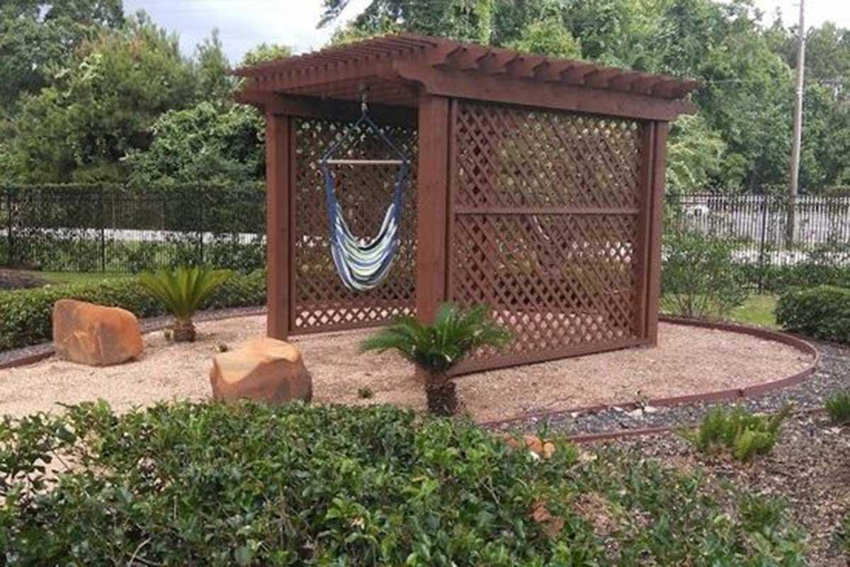 A wooden gazebo with a lattice design provides a shaded area for relaxation, featuring a colorful hammock hanging inside. Surrounding the gazebo are landscaped gardens with rocks and small plants, creating an inviting outdoor space in a tranquil setting.
