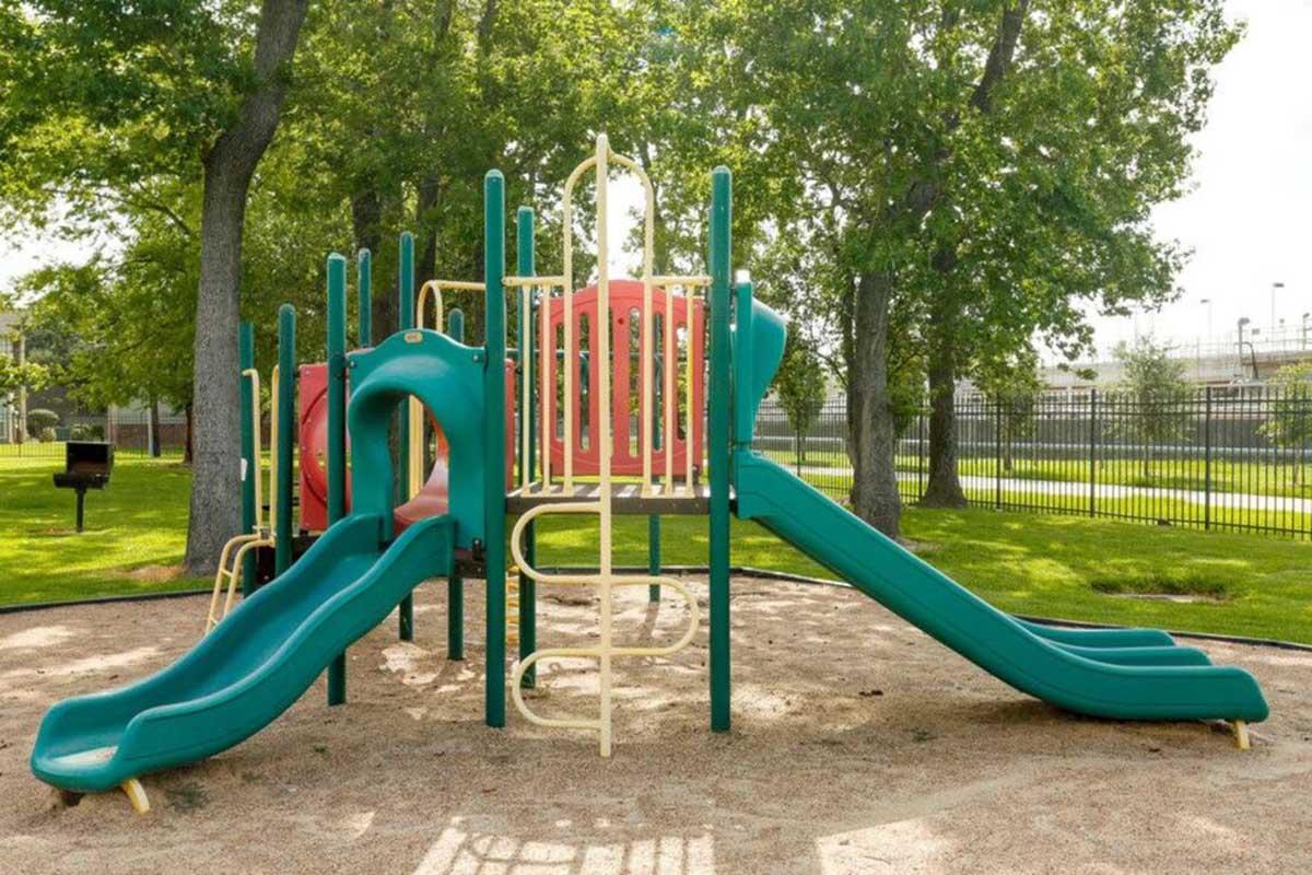 Colorful playground equipment featuring two slides, a climbing area, and a ladder, surrounded by green trees and grass. The playground is situated on sandy ground, creating a fun and safe environment for children to play. A fence is visible in the background.