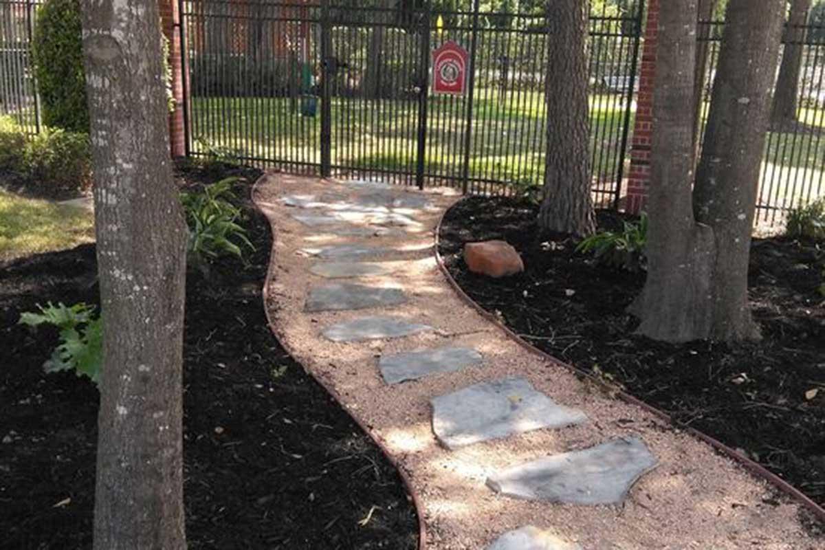 A winding pathway made of stone stepping stones, bordered by a gravel path, leads through a garden with lush greenery and trees. A metal fence encloses the area, providing a sense of privacy and security. The scene is serene, inviting exploration and nature appreciation.