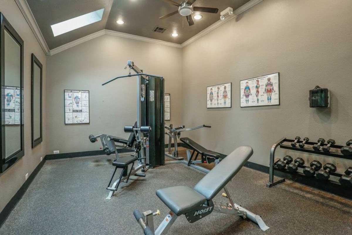 A small home gym featuring a multi-station workout machine, a weight bench, dumbbells on a rack, and fitness posters on the walls. The space has a textured floor, overhead fan, and a skylight providing natural light.