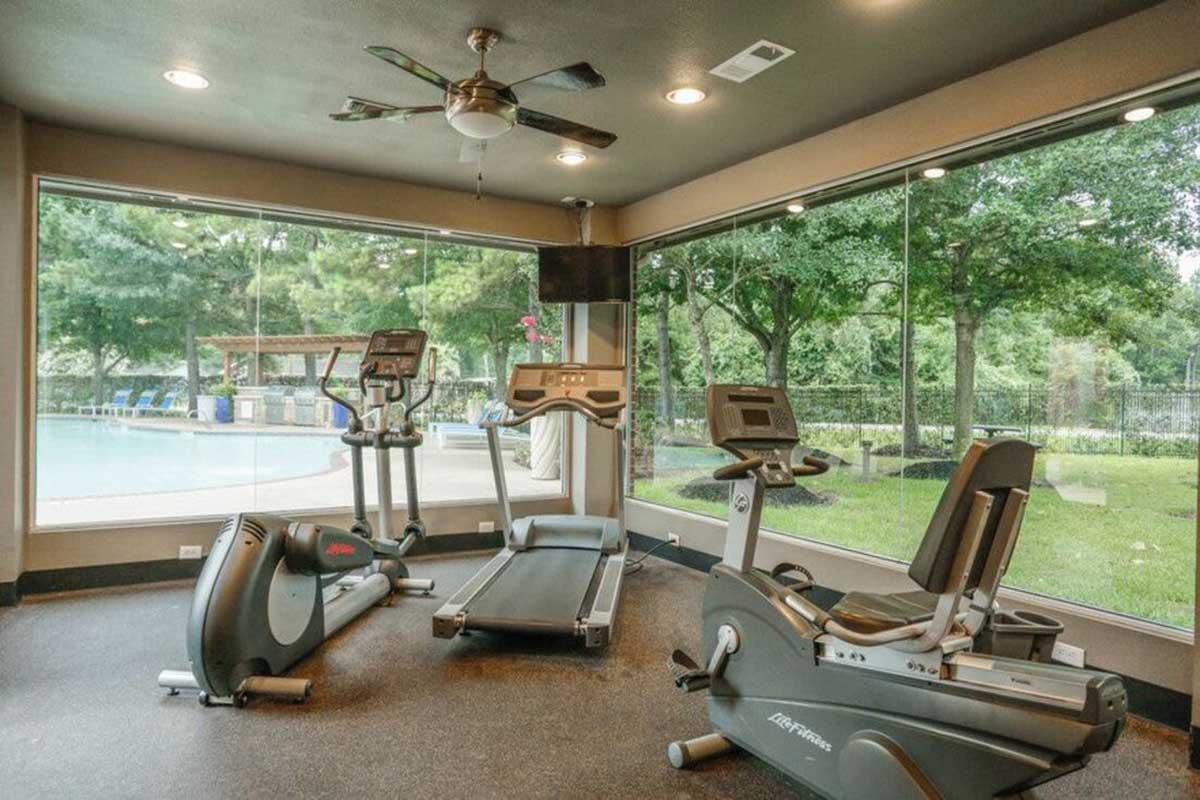 A modern home gym featuring a treadmill, elliptical machine, and stationary bike, with large windows showcasing a view of outdoor greenery and a swimming pool. The room is brightly lit with a ceiling fan and has a sleek, contemporary design.