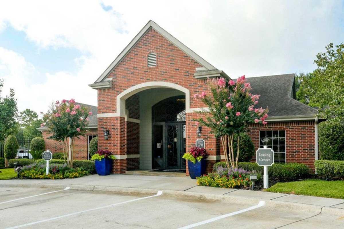 A brick building with a sloped roof and arched entrance, surrounded by colorful flowering plants and trees. The entrance features large windows and two blue planters. Signs indicate it is a "Leasing Center." The surrounding area includes a well-maintained lawn and parking spaces in front.