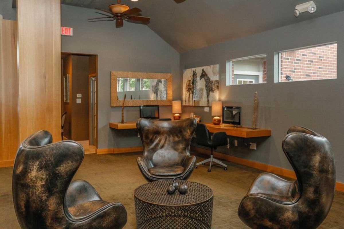 Cozy modern lounge area featuring two unique brown chairs with a textured finish, a round coffee table, and a wall-mounted mirror. The space is illuminated by soft lighting and includes a small workspace with a desk and computer. Large windows allow natural light to fill the room, creating an inviting atmosphere.