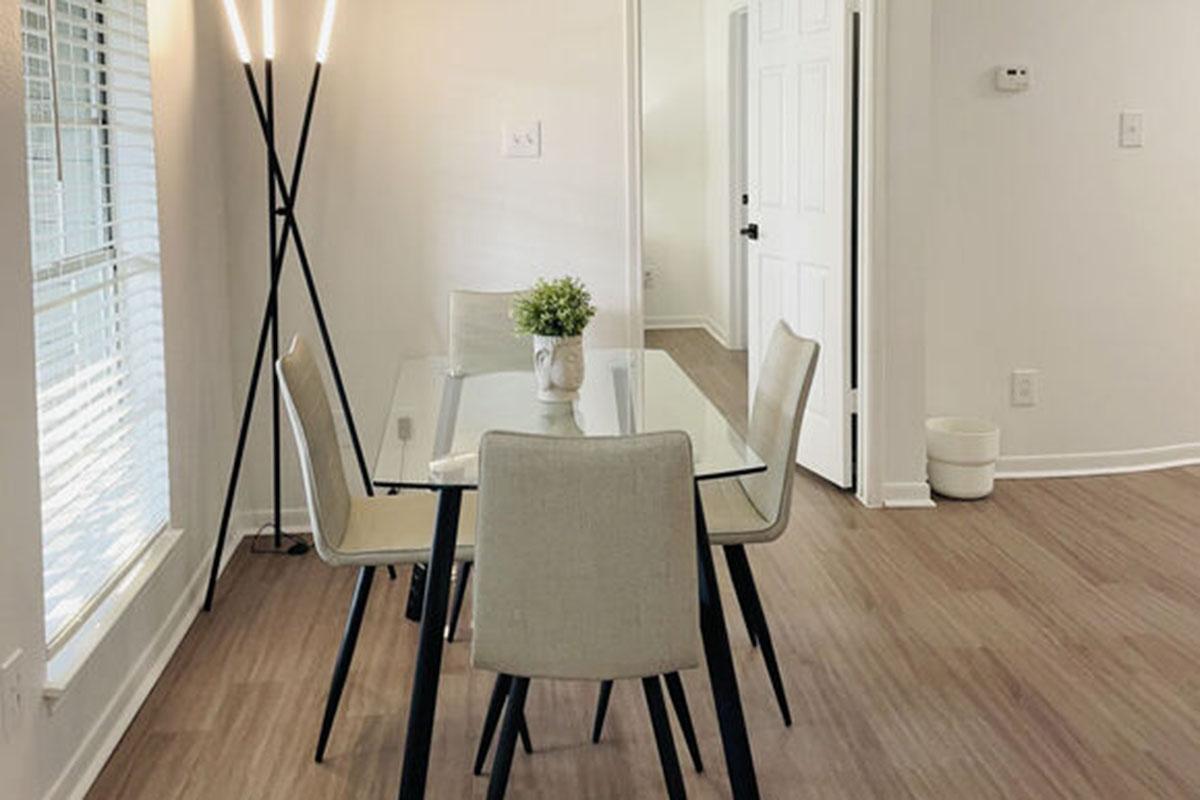 A modern dining area featuring a glass table with four beige chairs, a small plant centerpiece, and a stylish floor lamp. The room has hardwood flooring and bright natural light streaming through windows, creating an inviting and airy atmosphere. A door leads to another room.