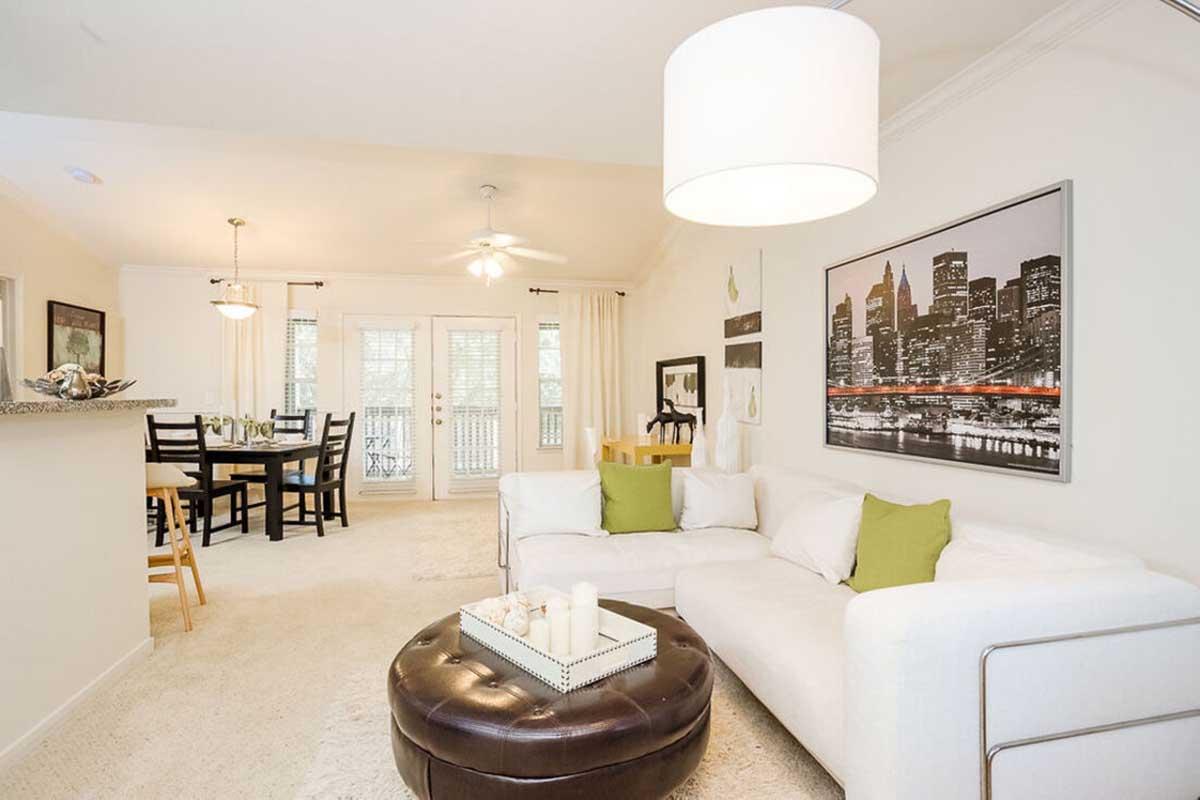 A cozy living room featuring a white sectional sofa with green pillows, a round coffee table, and a stylish lamp. The room has light walls, a fan, and a view of an outdoor balcony through sliding glass doors. A dining area with a wooden table and chairs is visible in the background, along with urban skyline artwork on the wall.