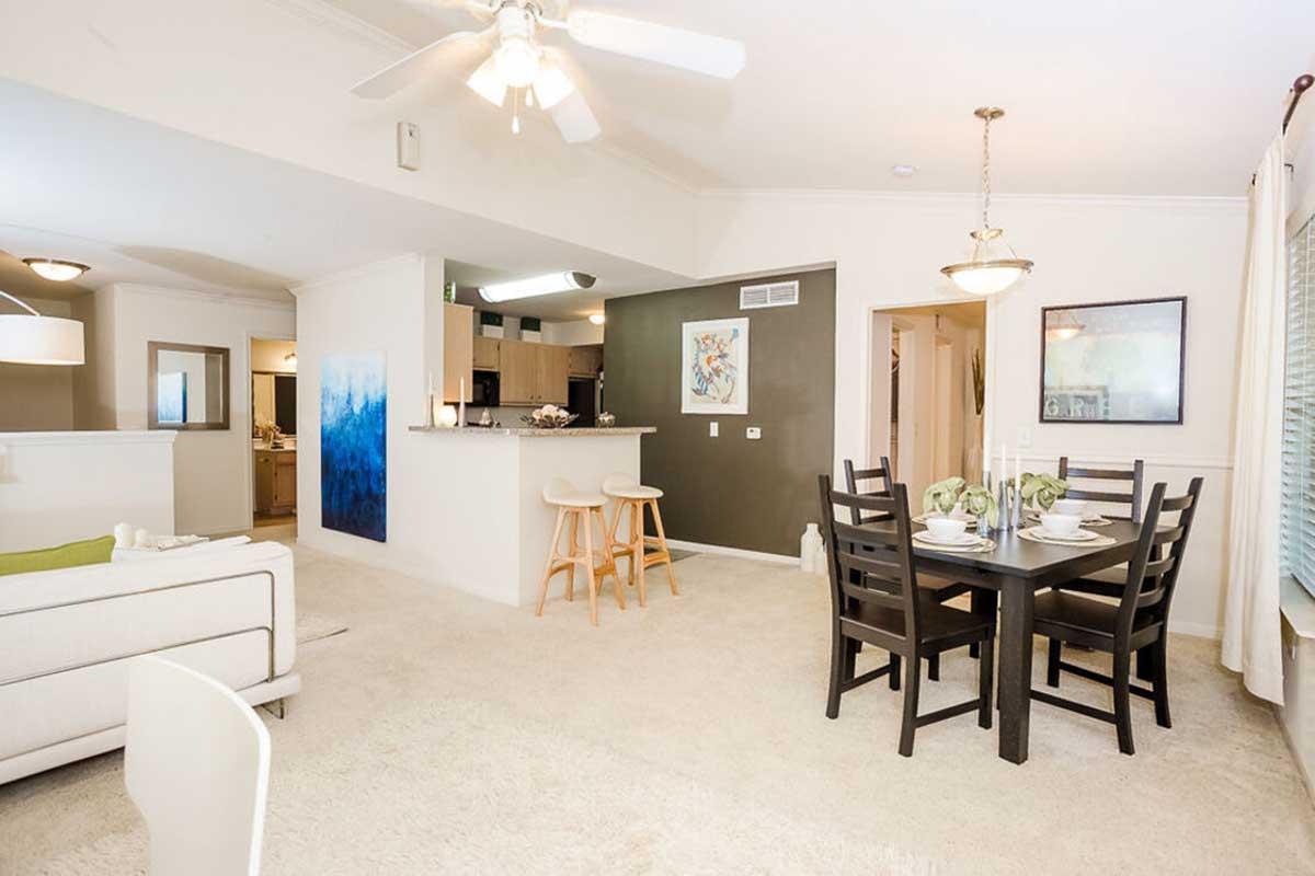 A bright, modern living space featuring a dining area with a dark wood table set for four. In the background, a kitchen with bar seating and a light-colored couch in the living room. Soft lighting and neutral walls create a warm atmosphere.