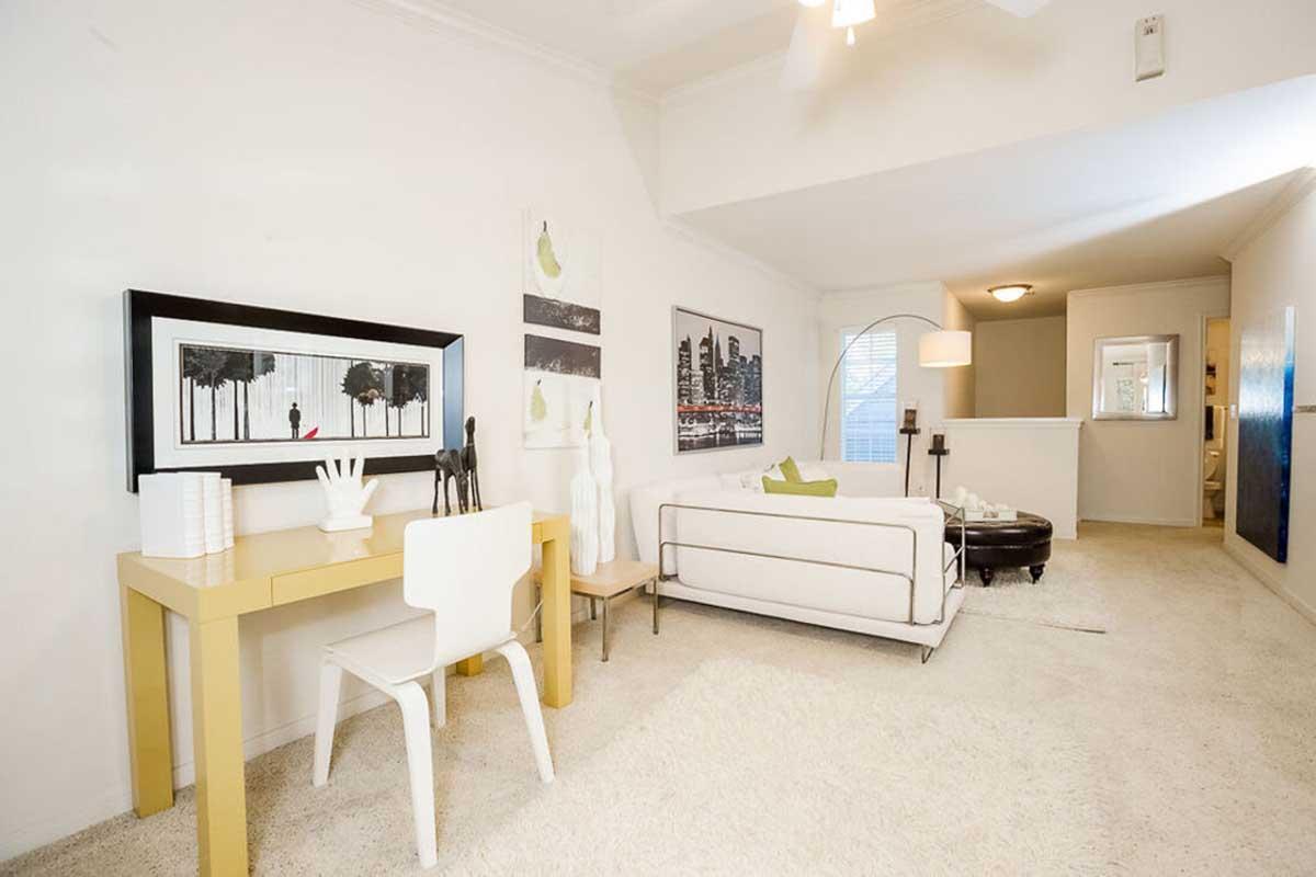A modern living room featuring a white sofa, a stylish coffee table, and a small desk with a chair. The decor includes framed artwork on the walls, a light color scheme, and a cozy carpet, creating a bright and inviting atmosphere.
