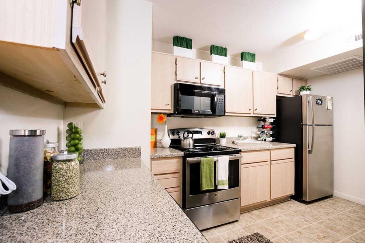Modern kitchen featuring light wooden cabinetry, a black microwave and oven, a stainless steel refrigerator, and granite countertops. Decorated with green accents, a potted plant, and kitchen accessories. The layout is open and bright with tiled flooring.