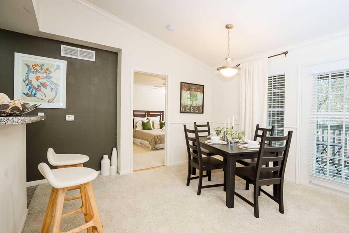 A modern dining area featuring a dark wooden table set for four, with white plates and green accents. In the background, a doorway leads to a bedroom. Light walls contrast with a dark accent wall, and large windows provide natural light. Stylish bar stools are positioned at a counter, enhancing the inviting atmosphere.