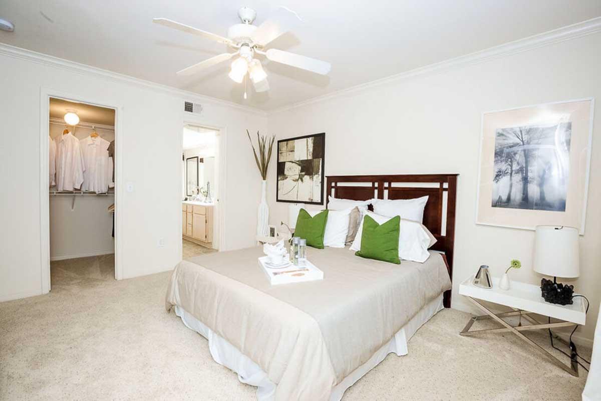 A cozy bedroom featuring a queen-sized bed with beige bedding and green accent pillows. A bedside table holds a tray with drinks and decor. There's a modern lamp and artwork on the walls. The room has a ceiling fan and a closet visible, showcasing neatly hung clothing. The carpet is light and soft, enhancing the inviting atmosphere.