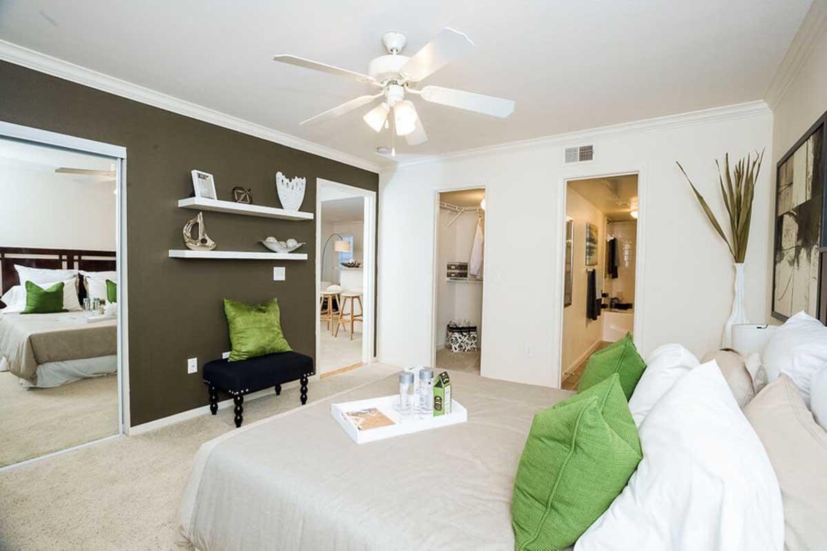 A cozy bedroom featuring a large bed with white pillows and green accents. A ceiling fan is overhead, and there's a small black bench at the foot of the bed. A shelf on the wall displays decorative items. Two doors lead to closets and a bathroom, with neutral-colored walls and soft carpet.