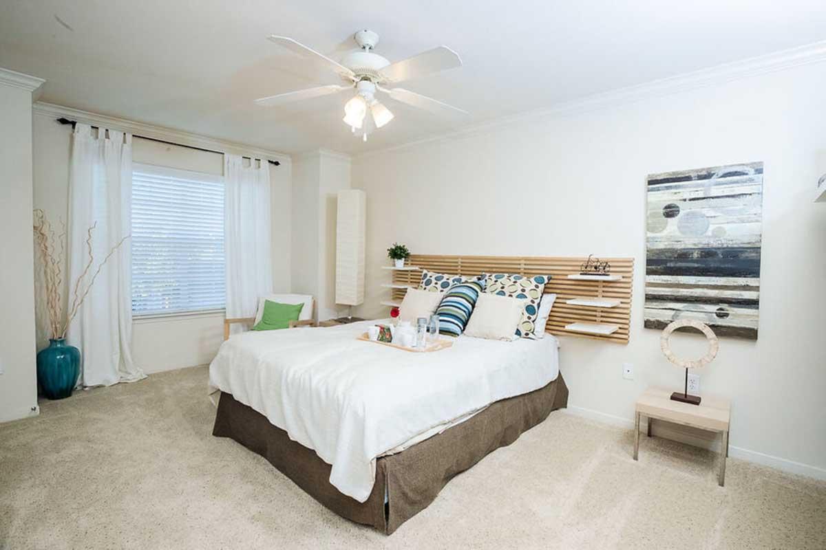A bright and airy bedroom featuring a bed with decorative pillows and a light-colored bedspread. There’s a wooden headboard with built-in shelves, a ceiling fan, and a window with sheer curtains. A decorative vase and a small bedside table add to the cozy ambiance. The walls are painted softly, enhancing the serene atmosphere.
