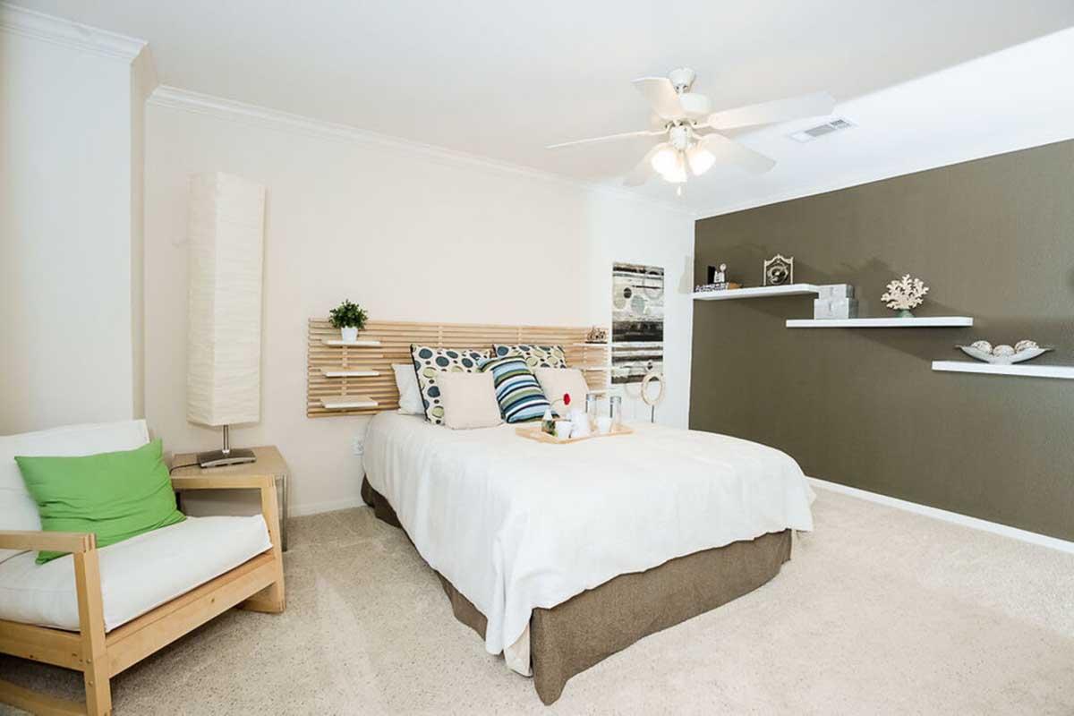A modern bedroom featuring a queen-sized bed with a wooden headboard, decorative pillows, and a bedside lamp. A light-colored armchair with a green cushion is beside the bed. The walls are painted in two colors: a light hue and a dark olive tone. A ceiling fan is above, and shelves with decorative items are mounted on the wall.
