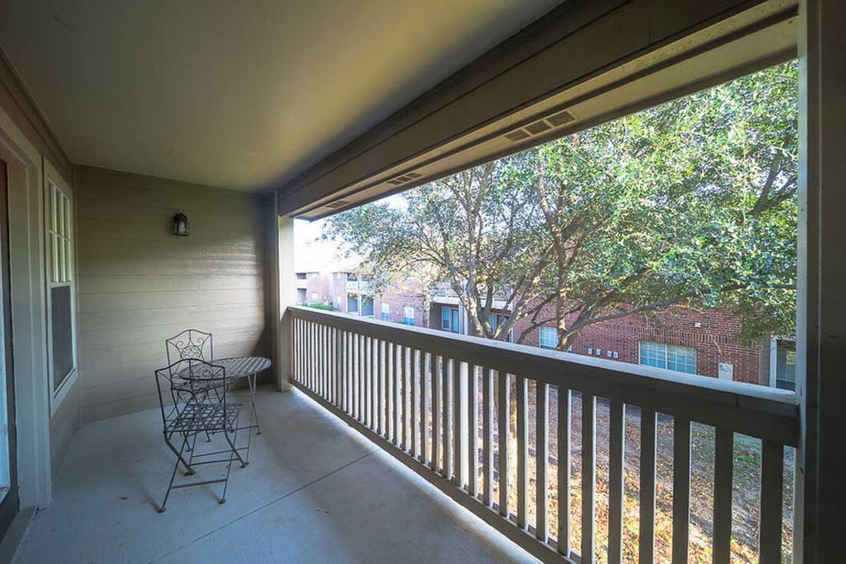 A small balcony view featuring a metal chair and table set. The railing provides a barrier, and lush green trees and nearby buildings are visible in the background. The atmosphere suggests a quiet, relaxing outdoor space.