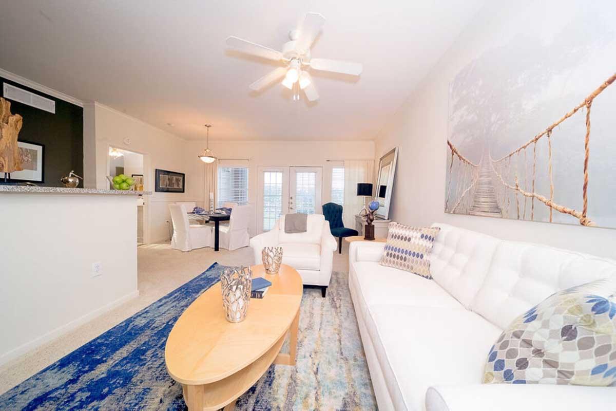 A modern living room featuring a white sofa with colorful throw pillows, a wooden oval coffee table, a blue area rug, and a ceiling fan. In the background, there’s a dining area with a dark table and chairs, and large windows allowing natural light to fill the space.