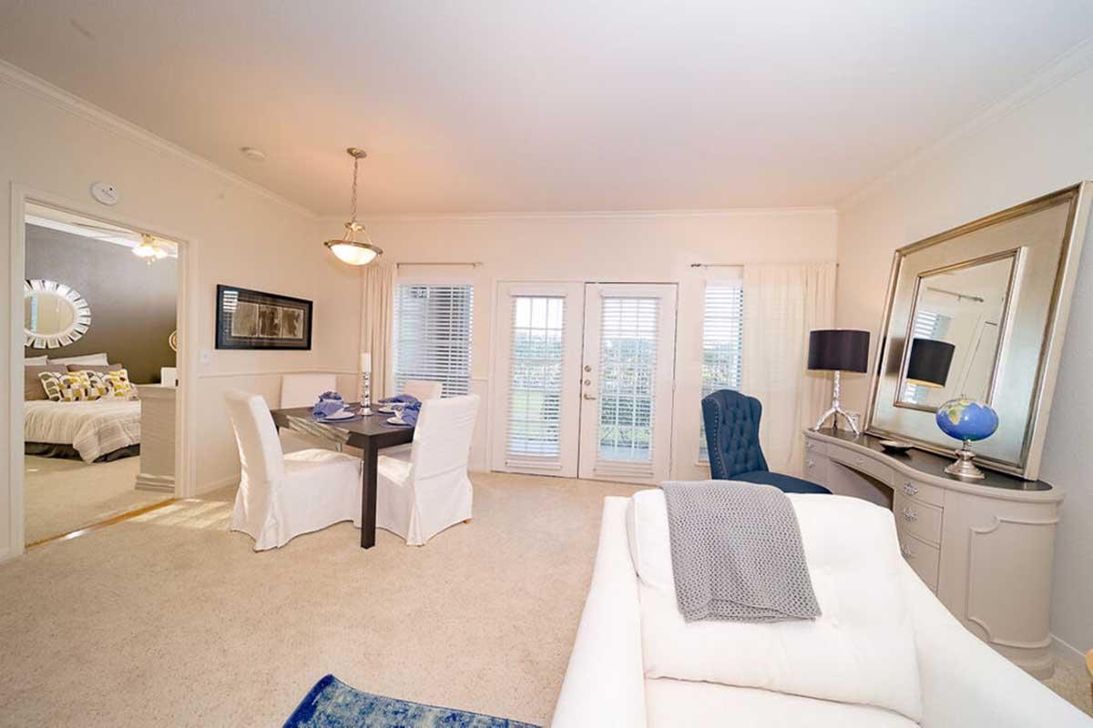 Cozy living space featuring a dining area with a round table and four chairs, a plush blue armchair, and a stylish console table with a lamp and globe. Bright double doors lead to a balcony, while light-colored walls and carpet give a warm, inviting atmosphere.