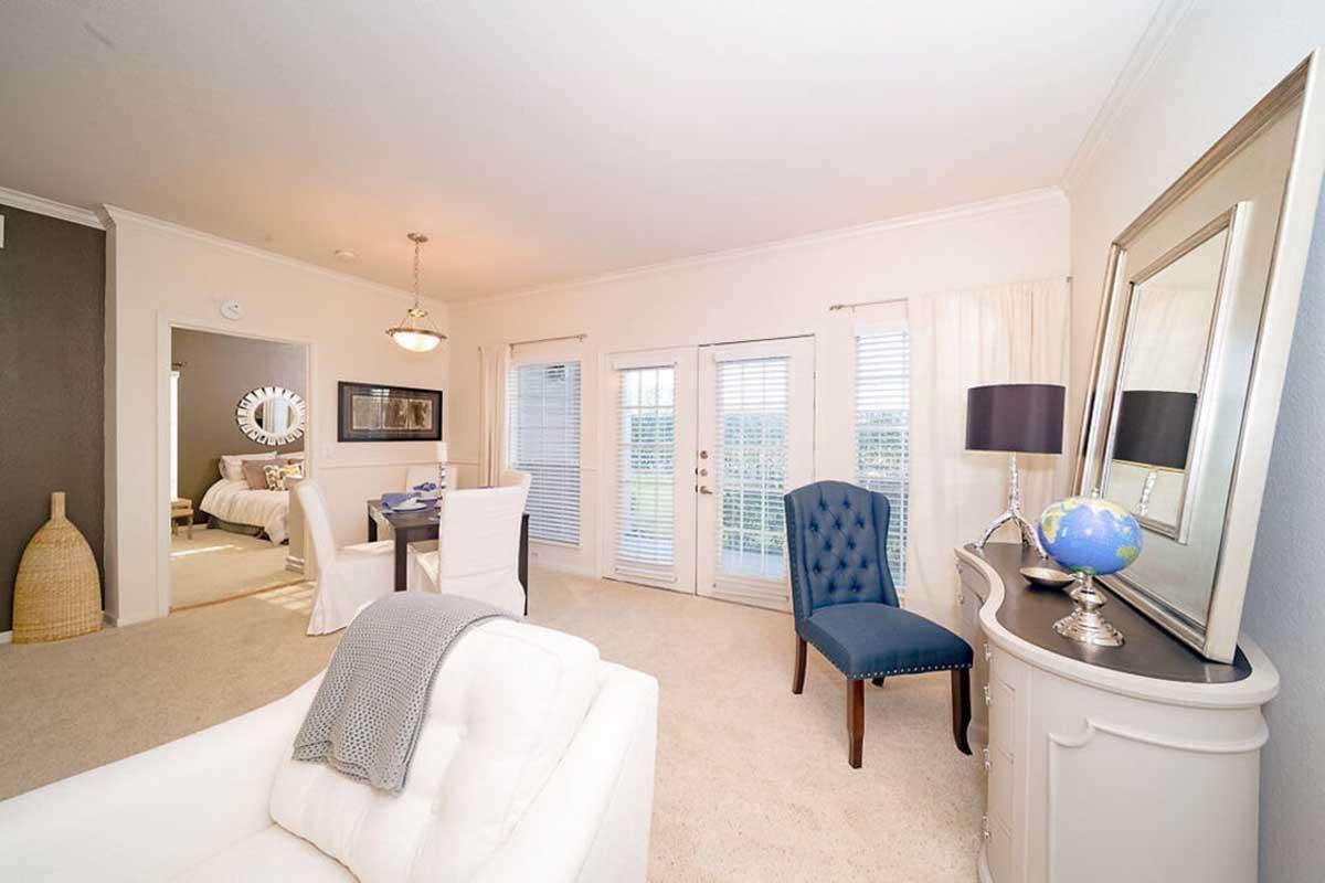 A well-lit living room featuring a white sofa, blue accent chair, and a decorative lamp on a table. Doors leading to a balcony are visible, alongside a dining area with a table set for four. The decor includes neutral tones and simple artwork, creating a cozy, inviting atmosphere.