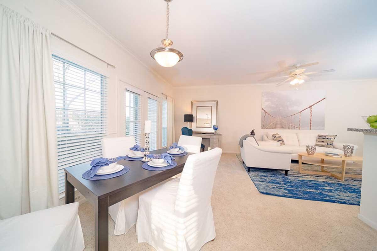 A bright dining area featuring a dark wooden table set with blue and white tableware. Surrounding the table are white chairs. The room has large windows with light-colored curtains, a light-colored sofa, and a blue area rug. A ceiling fan and wall art enhance the cozy atmosphere.