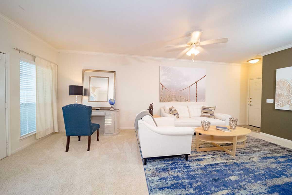 A bright, modern living room featuring a white sofa, a round coffee table, and a large area rug. There's a desk with a lamp and globe, a decorative mirror, and a wall with a scenic artwork. The space has a cozy, inviting ambiance with natural light from the windows.