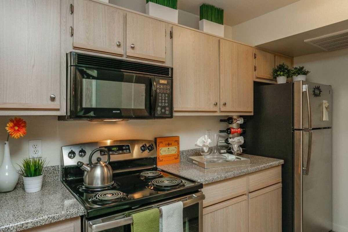 A modern kitchen featuring wooden cabinets, a black microwave above a stovetop, and a stainless steel refrigerator. The countertop is gray with a kettle, decorative items, and small plants. There's a pop of color with a flower vase on the countertop and a neatly organized kitchen space.