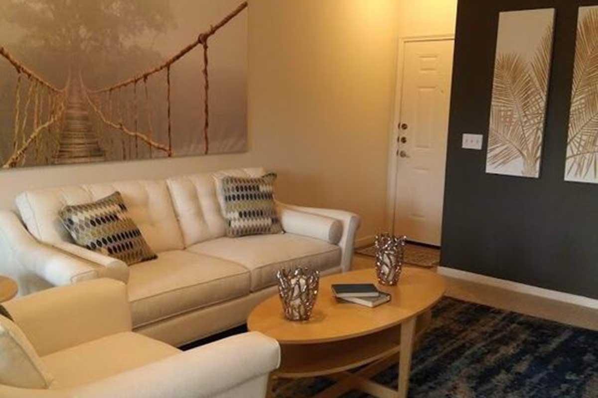 A cozy living room featuring a white sofa with patterned cushions, a round wooden coffee table with books, and decorative glass bowls. The wall displays a large nature-themed artwork of a bridge, and there are two framed prints of palm leaves on the adjacent wall. A doorway leads to another space.