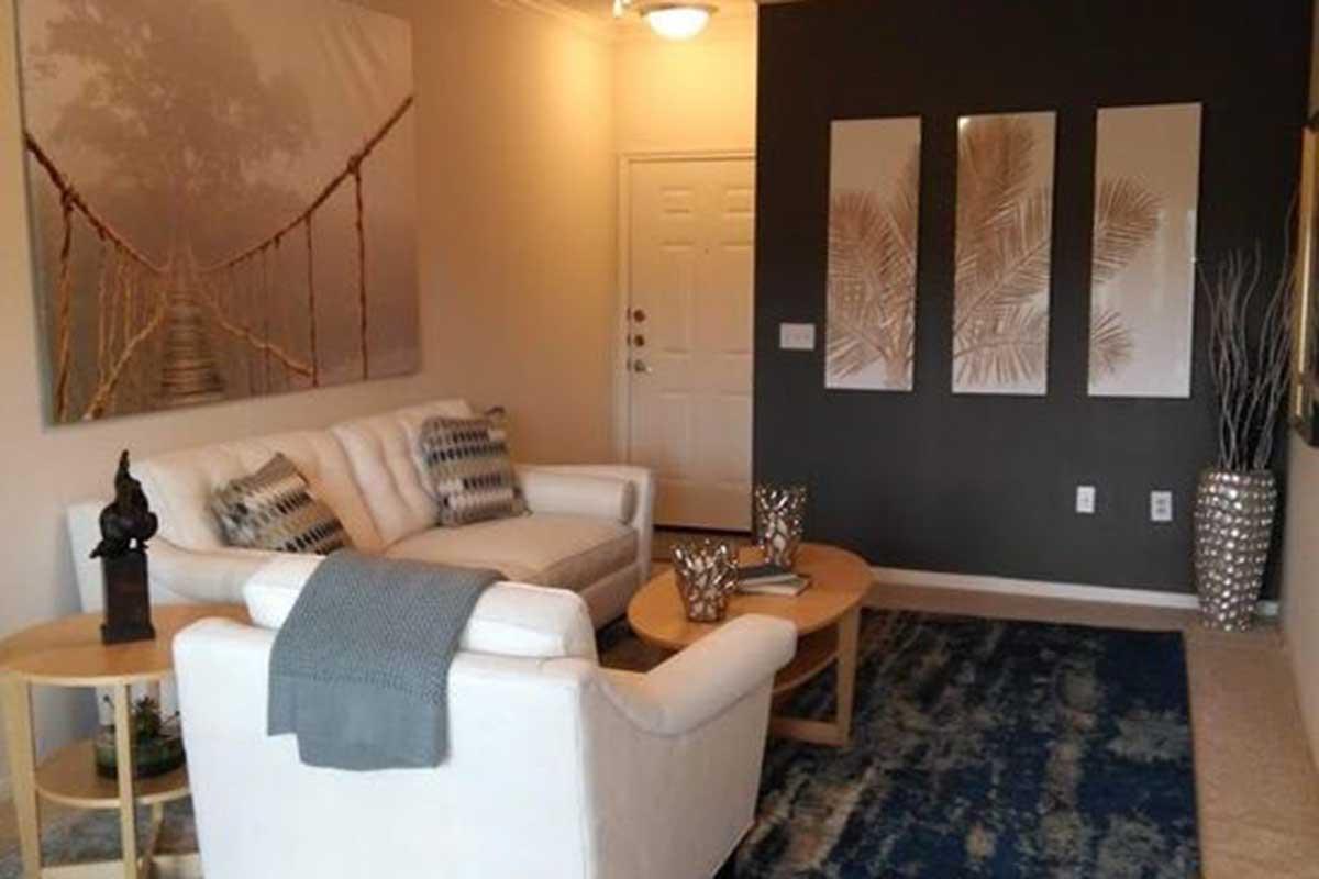 A cozy living room featuring a white sofa with decorative cushions, a round coffee table, and a textured blue rug. The walls are painted a dark color, and there is a large framed artwork of a foggy bridge. Three framed palm leaf designs hang above the sofa, and a small plant adds a touch of greenery.