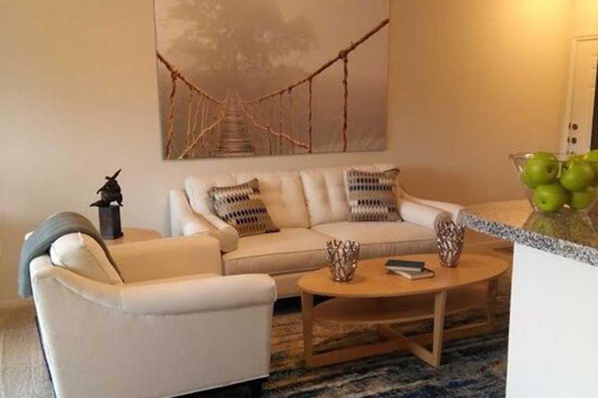 A cozy living room featuring a light-colored sofa and armchair, decorative pillows, a wooden coffee table, and a bowl of green apples. The wall above the sofa displays a large, serene landscape photograph of a bridge in misty surroundings. The space has a warm and inviting atmosphere.