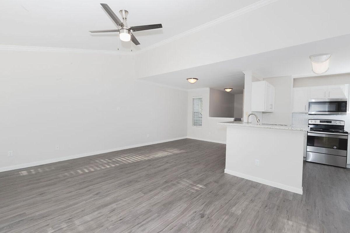 Spacious, modern living area featuring an open floor plan with light-colored walls, a ceiling fan, and hardwood flooring. A well-equipped kitchen is visible in the background, showcasing stainless steel appliances and a granite countertop, with ample natural light coming from nearby windows.