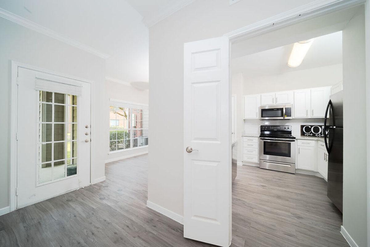 Interior view of a modern kitchen and adjacent room. The kitchen features white cabinetry, stainless steel appliances, and a light-colored countertop. There is a door leading to an outdoor area, and large windows allow for natural light to enter the space. The flooring is light laminate wood.