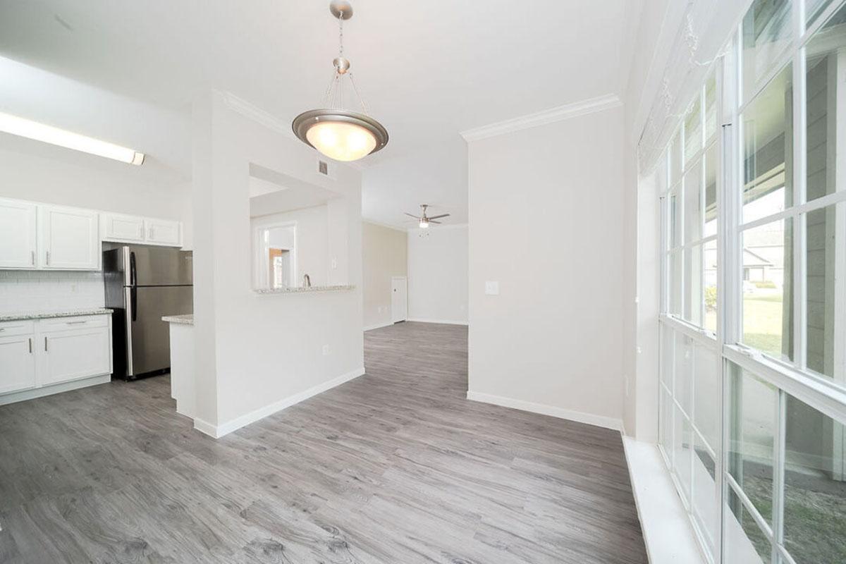 A bright, open living space featuring light gray flooring, white walls, and a modern kitchen with stainless steel appliances. A large window allows natural light to fill the area, and there is a ceiling fan for airflow. The layout connects the kitchen and living room, creating a spacious feel.