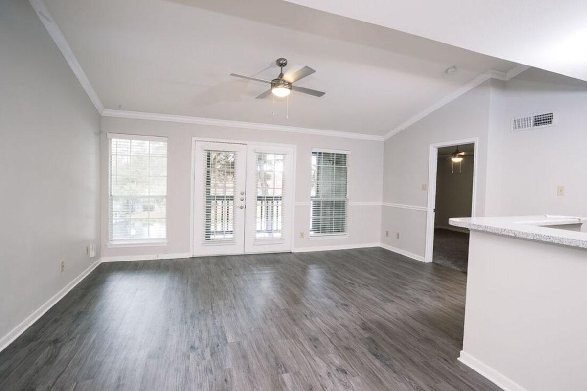 A spacious living room featuring a light gray color scheme, large windows with blinds, a ceiling fan, and a sliding glass door leading to a balcony. The flooring is wood-like laminate, and there is a partial view of an adjoining kitchen area in the background.